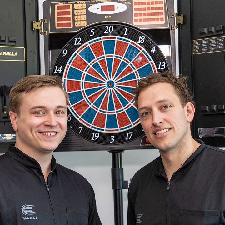 Zwei Männer stehen lächelnd vor einer elektronischen Dartscheibe. Beide tragen schwarze Jacken mit dem Logo „TARGET“.