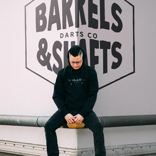 Barrels and Shafts Hoodie - Black Eine Person trägt einen schwarzen "Barrels and Shafts" Hoodie und sitzt auf einer Bank. Im Hintergrund ist ein großes Wandlogo mit der Aufschrift "Barrels & Shafts Darts Co" zu sehen.