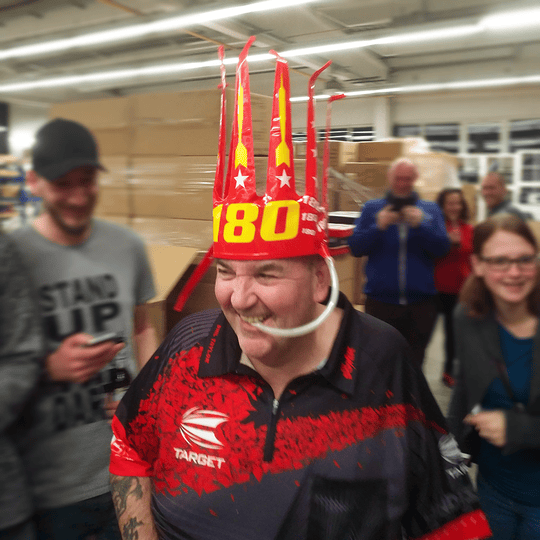 9026-McDart-180-er-Tr-tenkrone-4z2QvH75NbVwnP Ein lachender Mann trägt die "McDart 180-er Trötenkrone" auf dem Kopf. Um ihn herum stehen weitere Menschen und lachen ebenfalls.